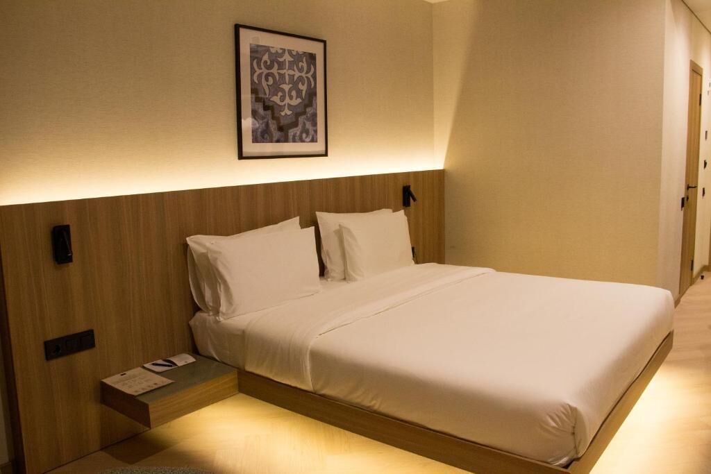 A hotel room with a bed, a bedside table, a framed picture on the wall, and a wooden headboard.