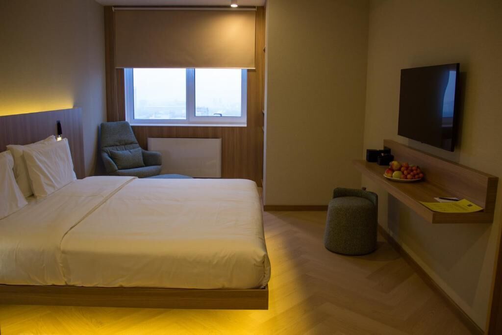 A hotel room with a bed, a chair, a TV, a small stool, and a window.