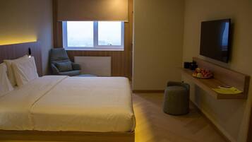 A hotel room with a bed, a chair, a TV, a small stool, and a window.