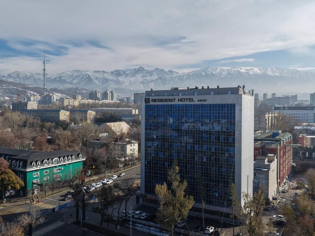 Un hôtel moderne à la façade vitrée, entouré de bâtiments urbains et avec une chaîne de montagnes en arrière-plan.