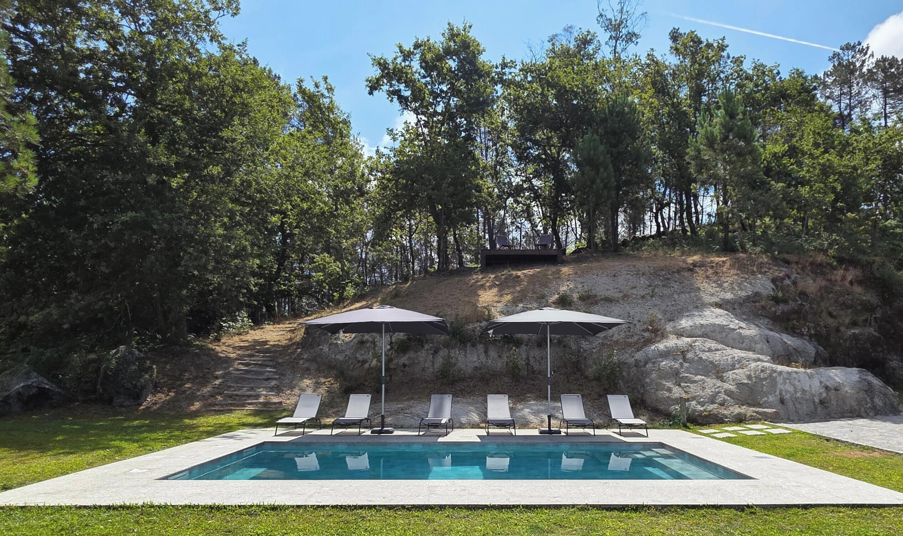 A swimming pool with lounge chairs and umbrellas, surrounded by trees and a rocky hillside.
