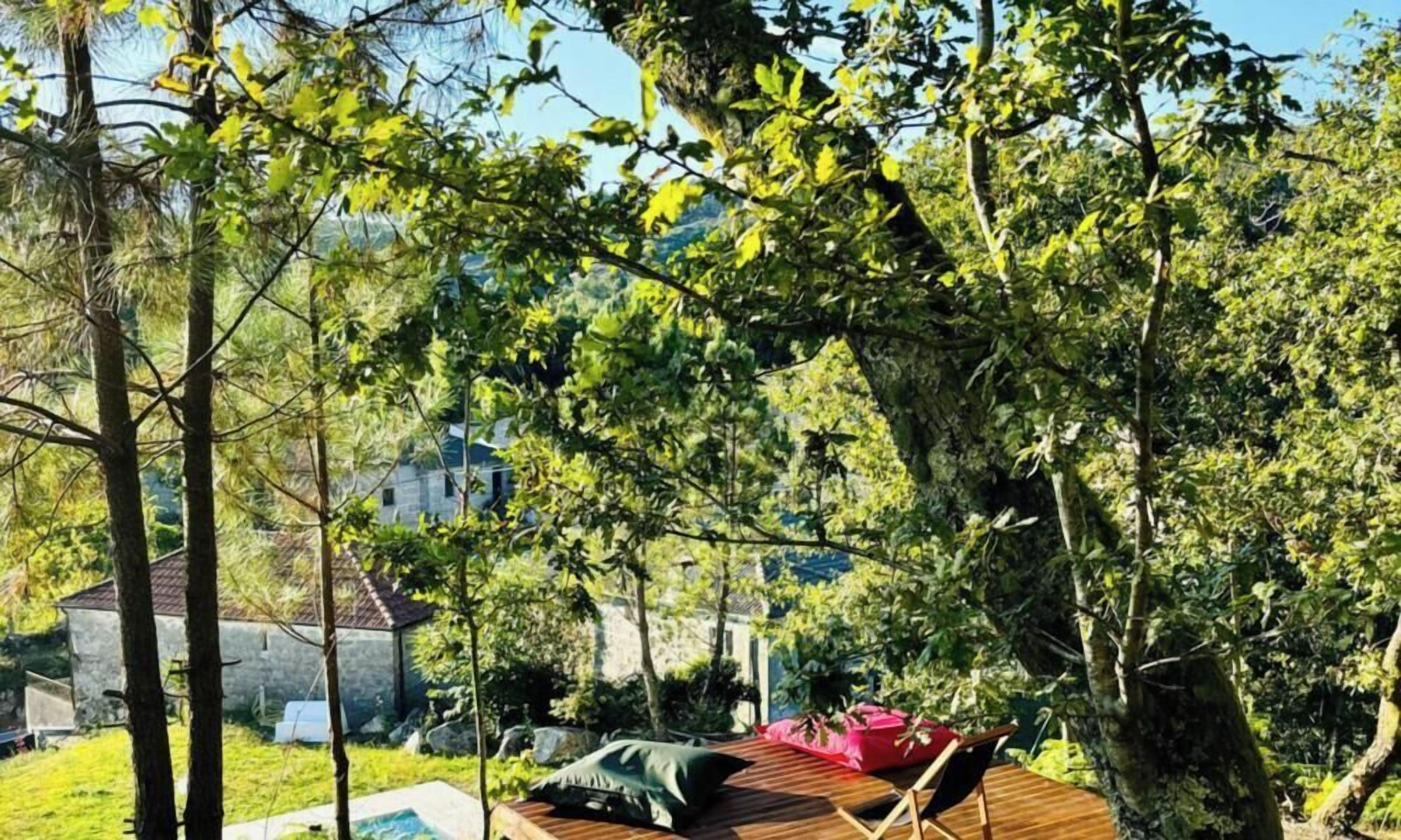 A wooden deck with a lounge chair and cushions, surrounded by trees and a stone building in the background.