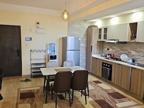 A compact kitchen with a refrigerator, stove, and sink. There is a dining area with a table and chairs. - Siaya Park - Elevate Stay (Kangundo)