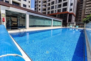 Una piscina con agua azul cristalina, rodeada por un edificio moderno con ventanales de vidrio y un techo de tejas.