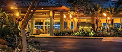 En hotellentrél med en skylt som visar texten ”MAHANA”, omgiven av tropiska växter och palmer.