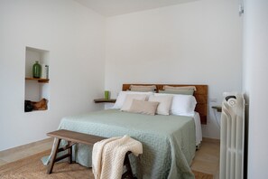 A bedroom with a bed, bedside tables, a radiator, and a small shelf with decorative items. - Beyond the Cones (Puglia)
