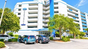 Et hotel i flere etager med et markant blå og hvidt farvetema, der viser en tydelig indgang og en parkeringsplads med flere biler.