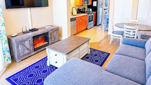 En moderne stue med sofa, sofabord og pejs. Et køkken med hårde hvidevarer samt et spiseområde med stole kan ses i baggrunden.