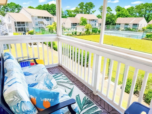 A balcony with a sofa, a dining table, and chairs. There is a view of a residential area with houses and a tennis court. - Little River Inn 1507 (Little River)