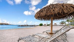 Ein Strand mit Liegestühlen und einem von Stroh bedeckten Sonnenschirm, ein klarer Himmel und Boote im Hintergrund.