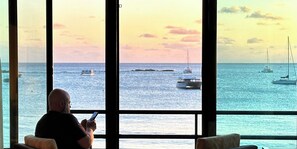 A man sitting by a large window overlooking a calm sea with boats.