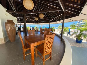 A covered outdoor dining area with wooden tables and chairs, a large vase, and a view of the beach and ocean.