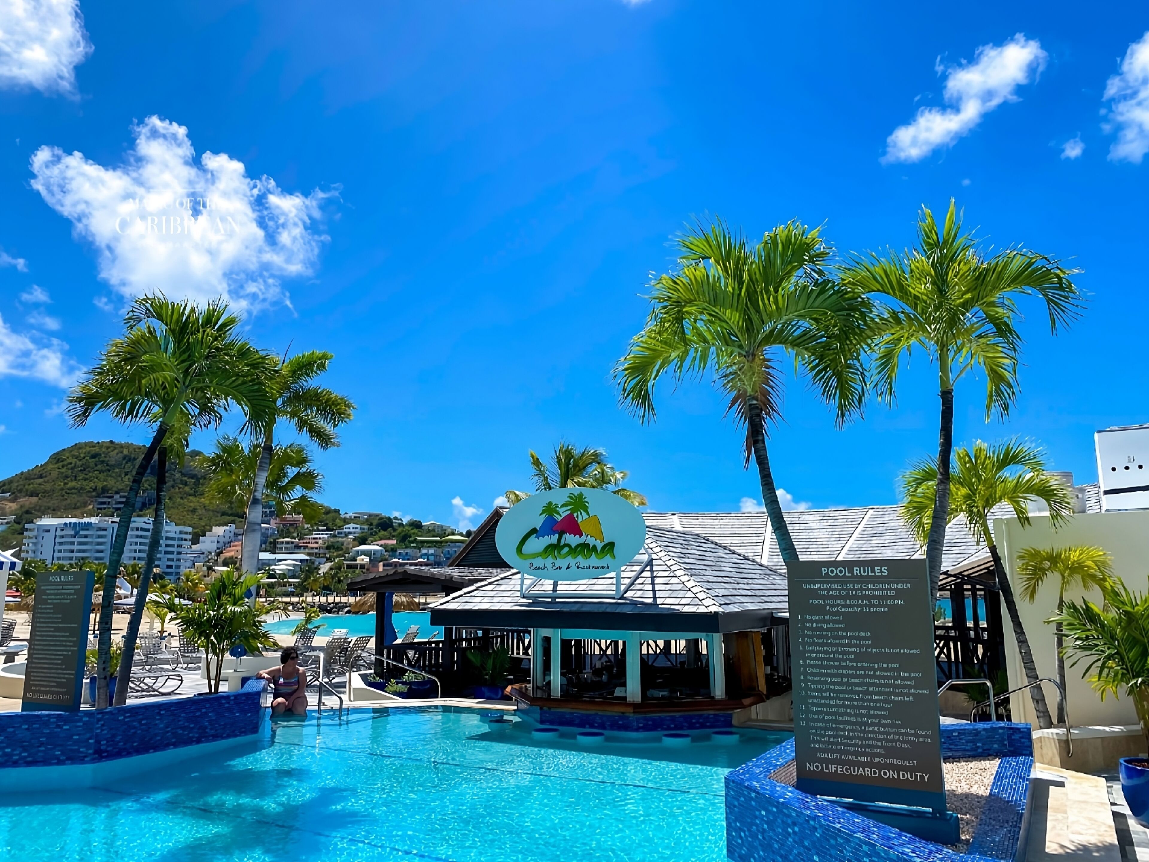 Una piscina azul cristalina rodeada de palmeras y un bar en la playa, con vistas a una colina al fondo.