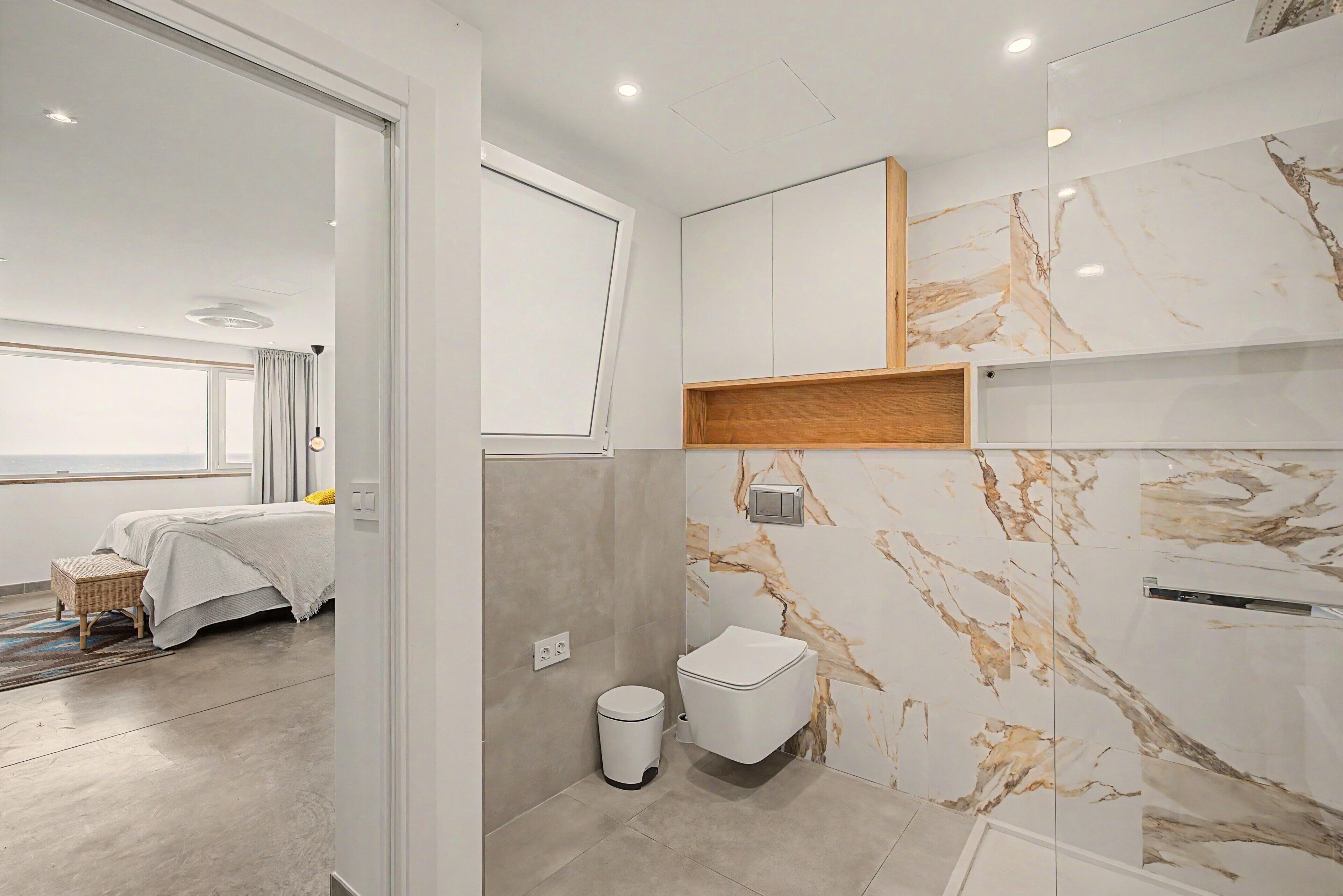 A modern bathroom with marble walls, a toilet, and a small trash bin.