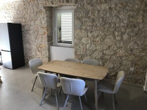 A stone-walled room with a wooden table and four chairs, a window with blinds, and a black cabinet.