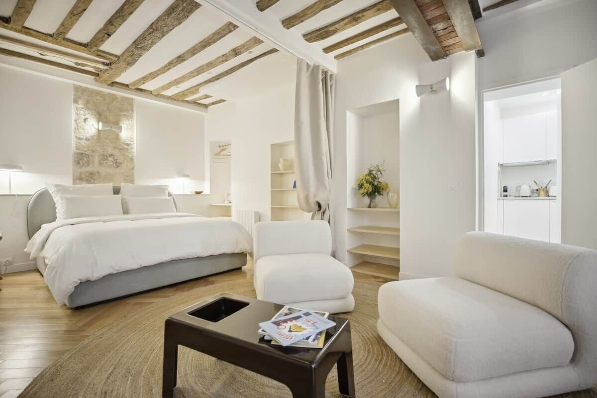 A modern hotel room with a wooden ceiling, a bed, a sofa, a coffee table, and a staircase leading to a kitchen area.