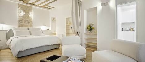A modern hotel room with a wooden ceiling, a bed, a sofa, a coffee table, and a staircase leading to a kitchen area.