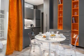 A modern kitchen with a dining area, featuring a round table with chairs, a refrigerator, and shelves with items. - Grand confort au pied du Louvre (Paris)