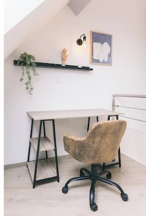 Ein Zimmer mit Schreibtisch, Stuhl, einer Pflanzenregal und einem Wandleuchte sowie einem gerahmten Bild.