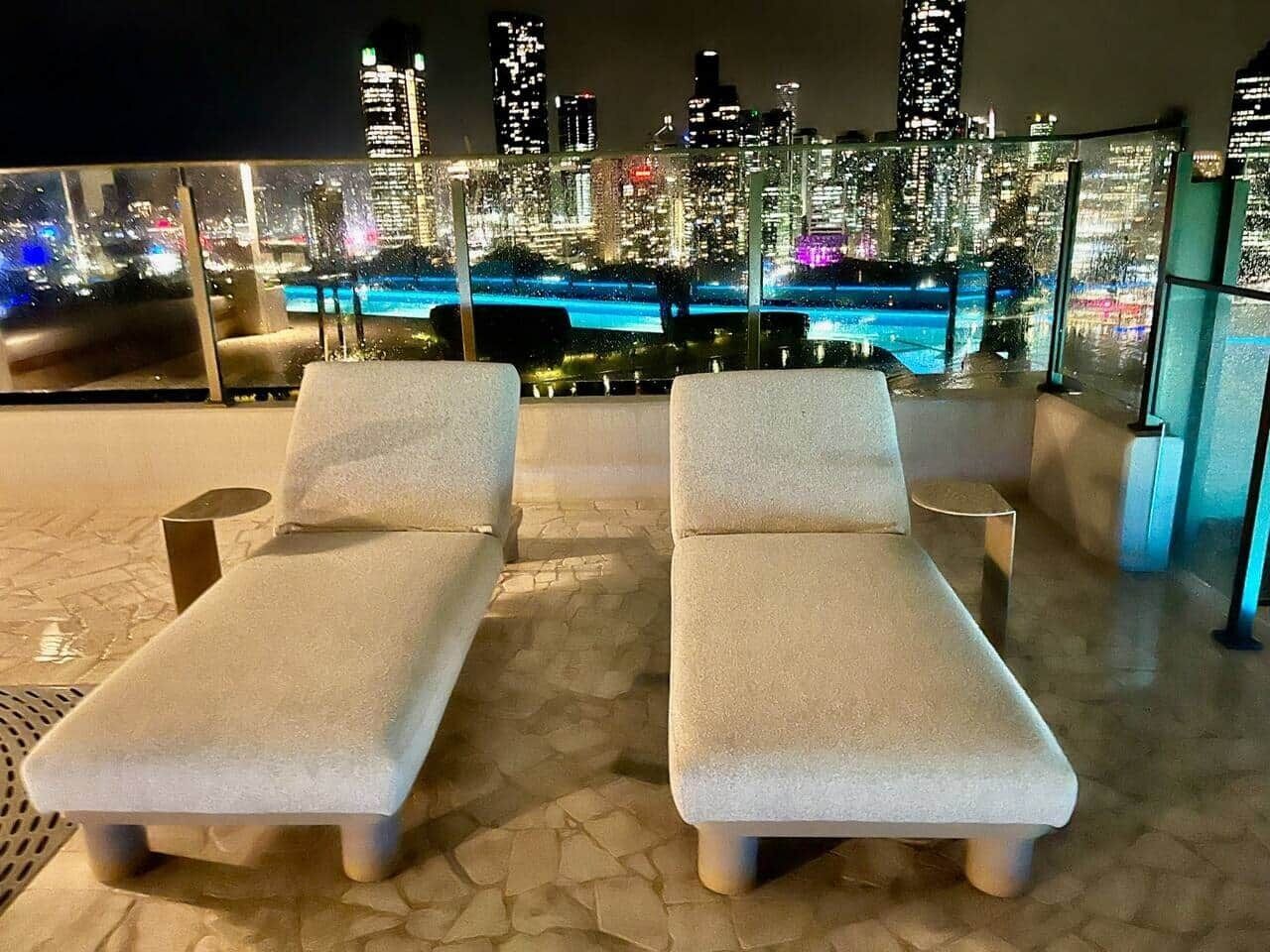 Two lounge chairs with cushions, a small table, and a cityscape view at night.