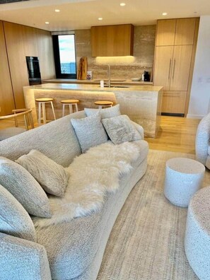 A modern living room with a sofa, ottoman, and a kitchen area with wooden cabinets and a bar. - Luxury 2BR Apt, Pool, Parking, Skye Kangaroo Pt (Kangaroo Point)
