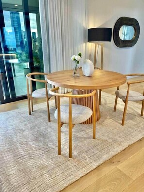A round wooden dining table with four chairs, a woven rug, a white vase with flowers, a black floor lamp, and a round mirror on the wall. - Luxury 2BR Apt, Pool, Parking, Skye Kangaroo Pt (Kangaroo Point)