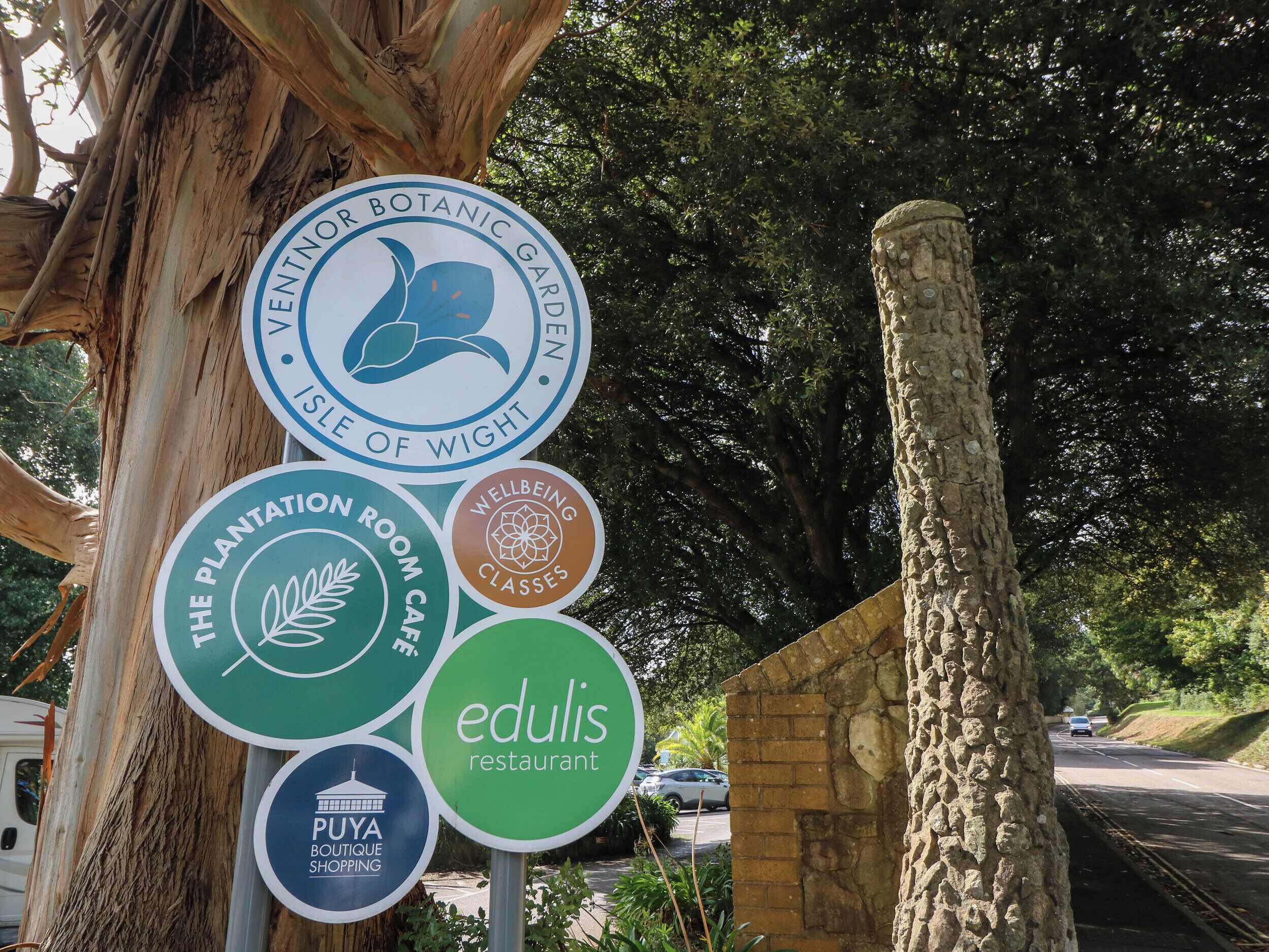 A sign with multiple circular logos indicating a botanical garden, a plantation room cafe, wellbeing classes, a restaurant named Edulis, and a boutique shopping area.