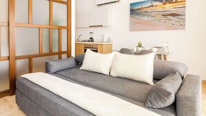 Quarto moderno com sofá-cama, uma kitchenette compacta e uma pintura de uma paisagem praiana.