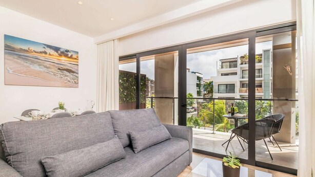 Uma sala de estar moderna com um sofá cinzento, uma mesa de jantar com cadeiras e uma grande janela com vista para a paisagem urbana.
