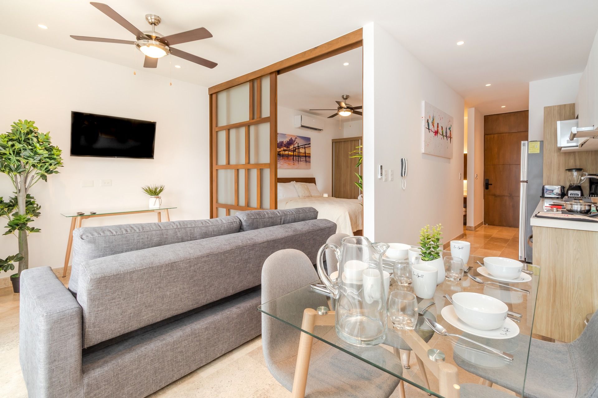 A modern living room with a grey sofa, a glass dining table set with white dishes, and a kitchen area with a refrigerator and a coffee machine.