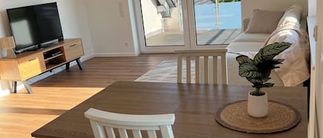 A modern living room with a wooden dining table, white chairs, a flat-screen TV, and a sofa.
