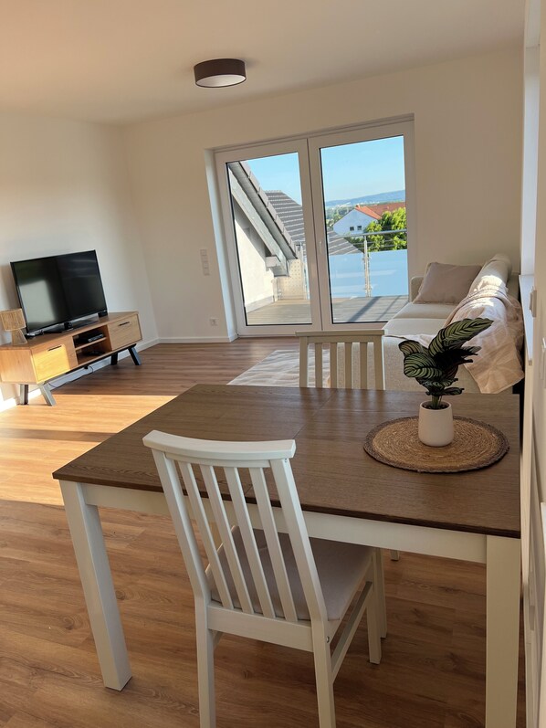 A modern living room with a wooden dining table, white chairs, a flat-screen TV, and a sofa.