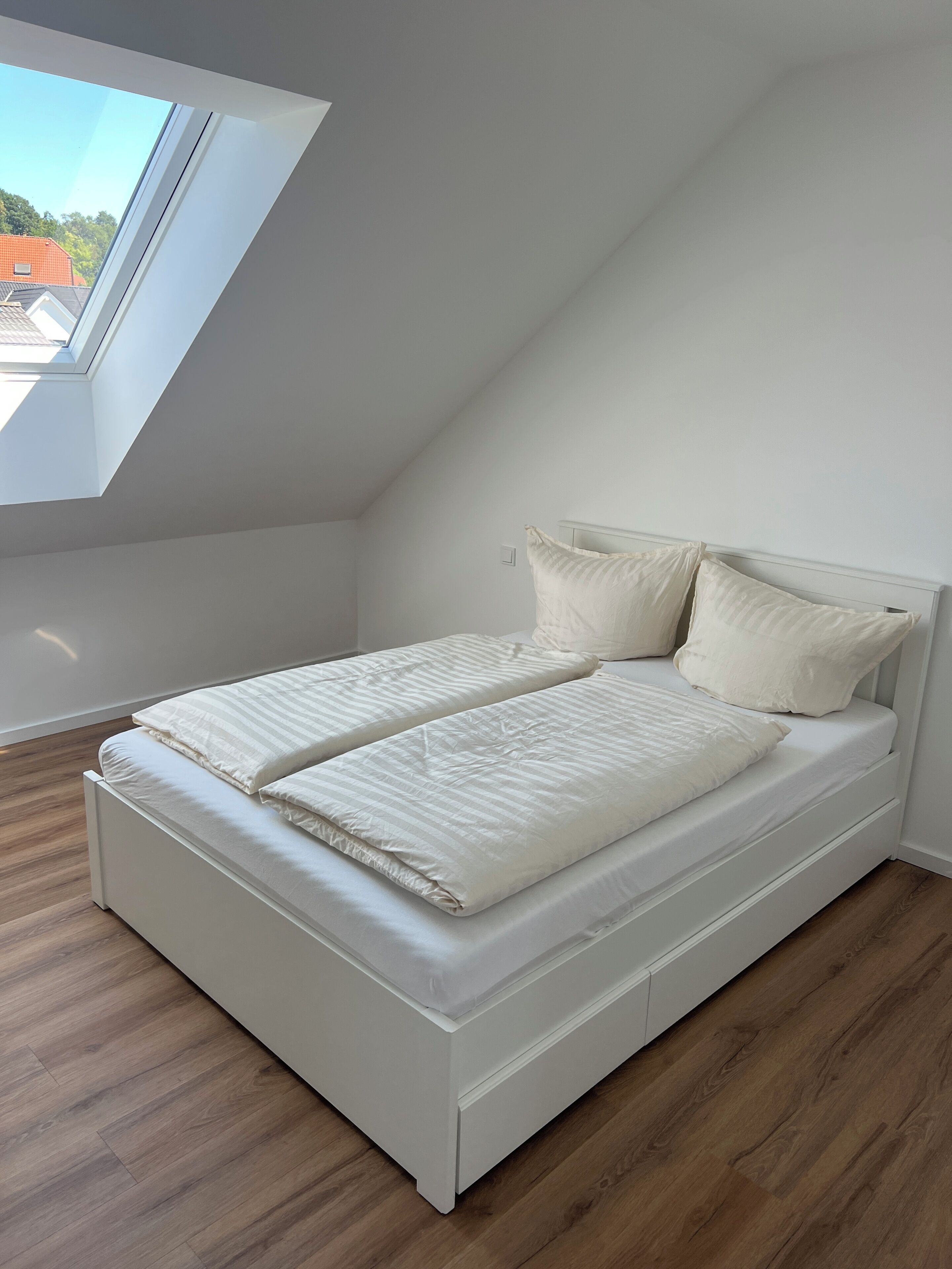 Quarto com teto inclinado, claraboia, cama com roupa de cama branca e soalho de madeira.