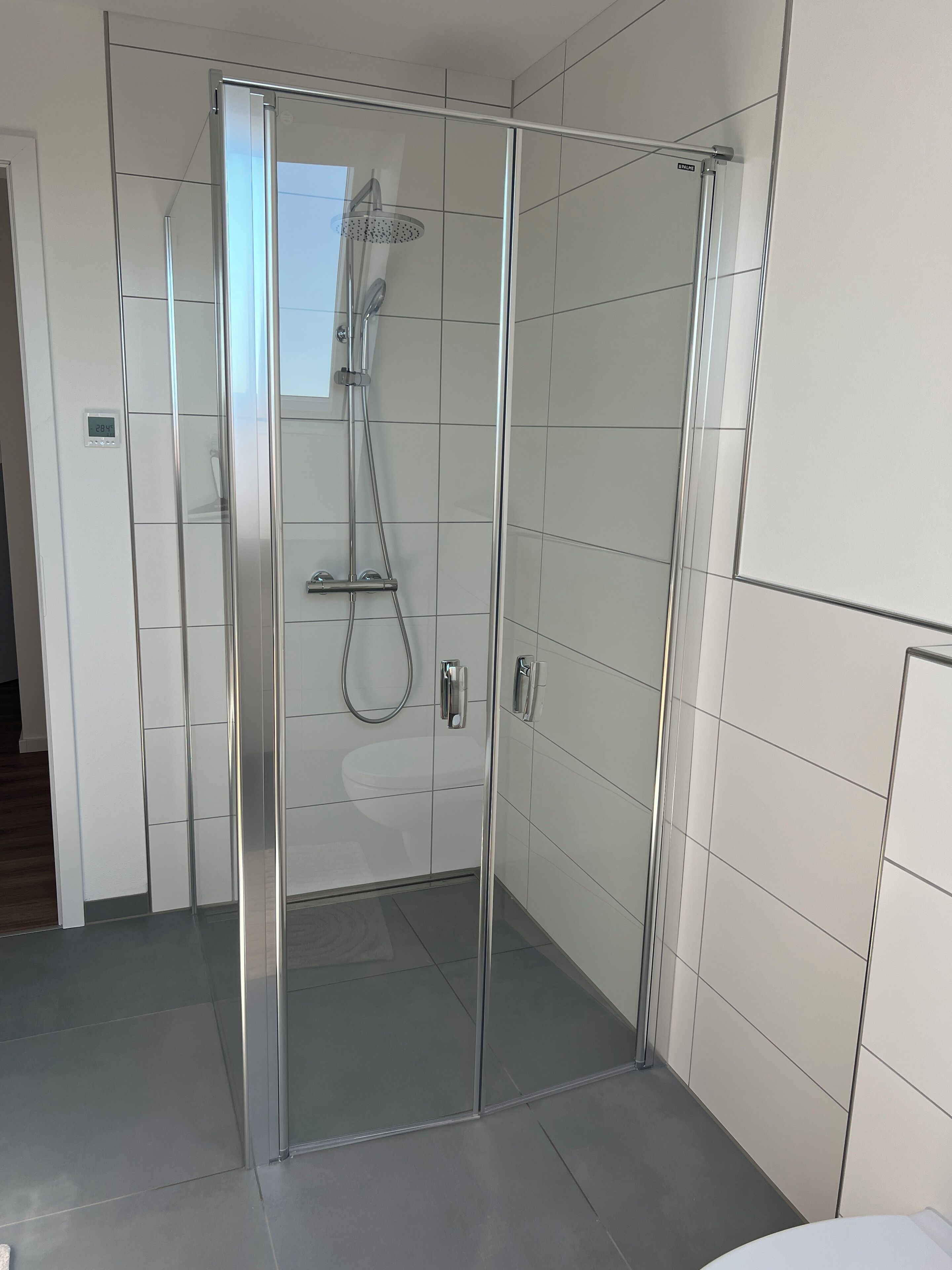 Ein modernes Badezimmer mit einer gläsernen Duschkabine, weißen Fliesen und einer sichtbaren Toilette.