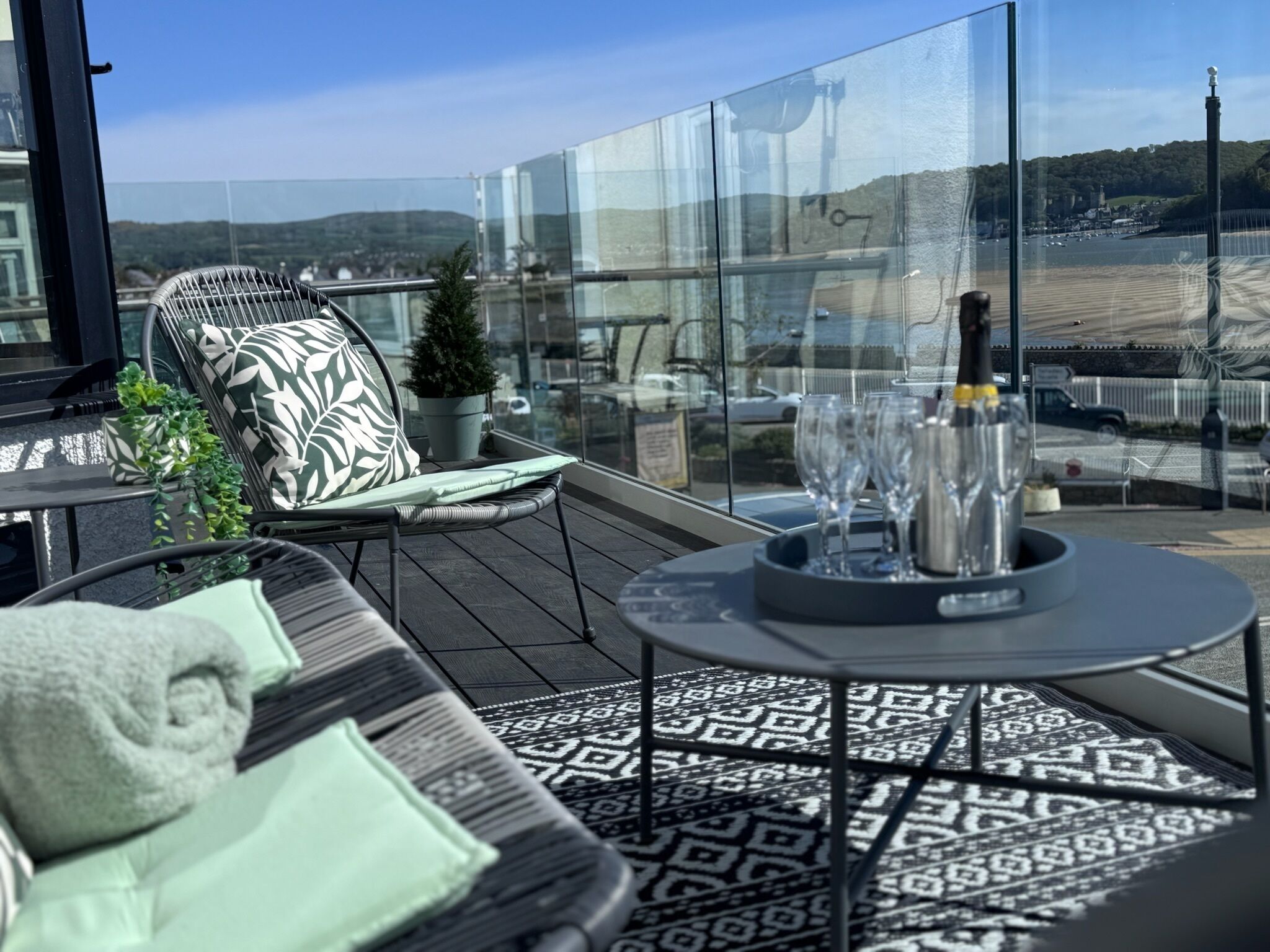 A balcony with a glass partition, a patterned floor, a cushioned chair, a small potted plant, and a table with wine glasses and a bottle.