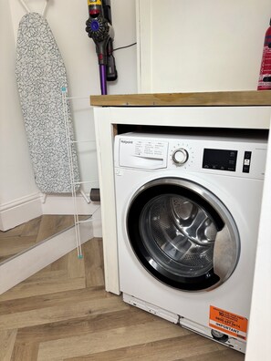 A compact washing machine with a control panel, a wooden shelf above it, and a hanging ironing board. - Stunning family friendly Apartment with Balcony & Views (Deganwy)