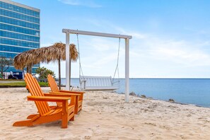 Une plage avec une chaise suspendue et deux chaises Adirondack, un parasol en chaume et, au loin, un bâtiment moderne.