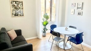 A modern living room with a dark sofa, a round dining table with blue chairs, a potted plant, and a window with curtains. - Rooms with a View, Trastevere Home Rental (Roma)
