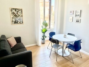 Ein modernes Wohnzimmer mit einem dunklen Sofa, einem runden Esstisch mit blauen Stühlen, einer Topfpflanze und einem Fenster mit Vorhängen.