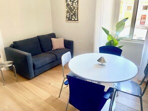 Ein modernes Wohnzimmer mit einem dunkelblauen Sofa, einem gemusterten Kissen, einem runden weißen Tisch mit einer Lampe und einer Topfpflanze.