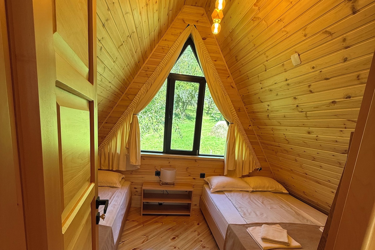 Acogedor dormitorio en ático con paredes de madera y techo inclinado, con dos camas y una ventana con vista a follaje.