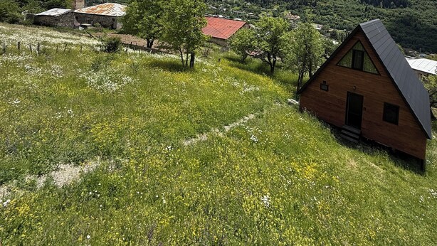 Una cabaña de madera con techo inclinado, rodeada por un prado verde exuberante lleno de flores silvestres, con montañas y un cielo parcialmente nublado al fondo.