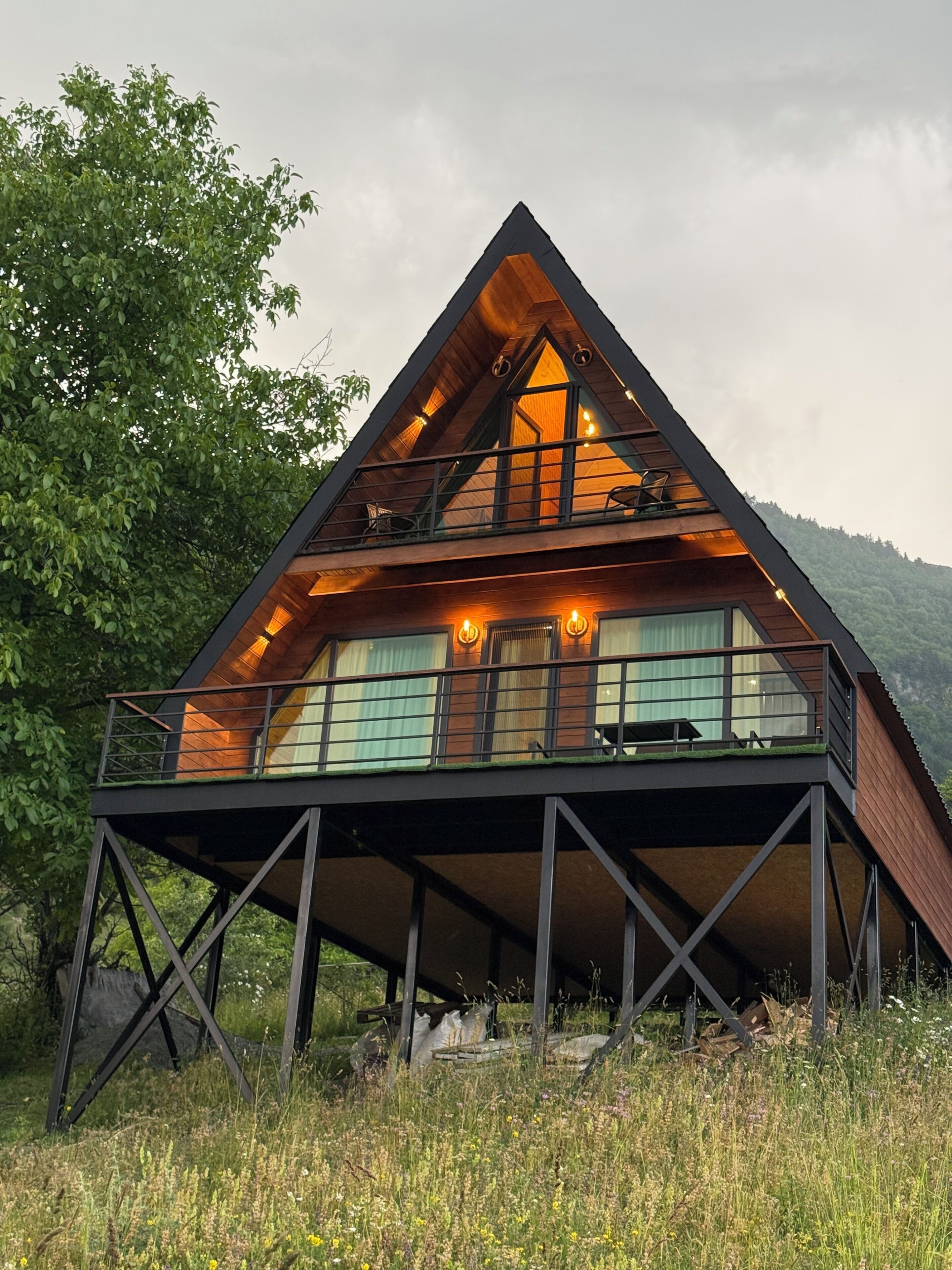 Une cabane moderne en bois, de forme triangulaire, dotée d’un balcon et entourée d’un paysage naturel.