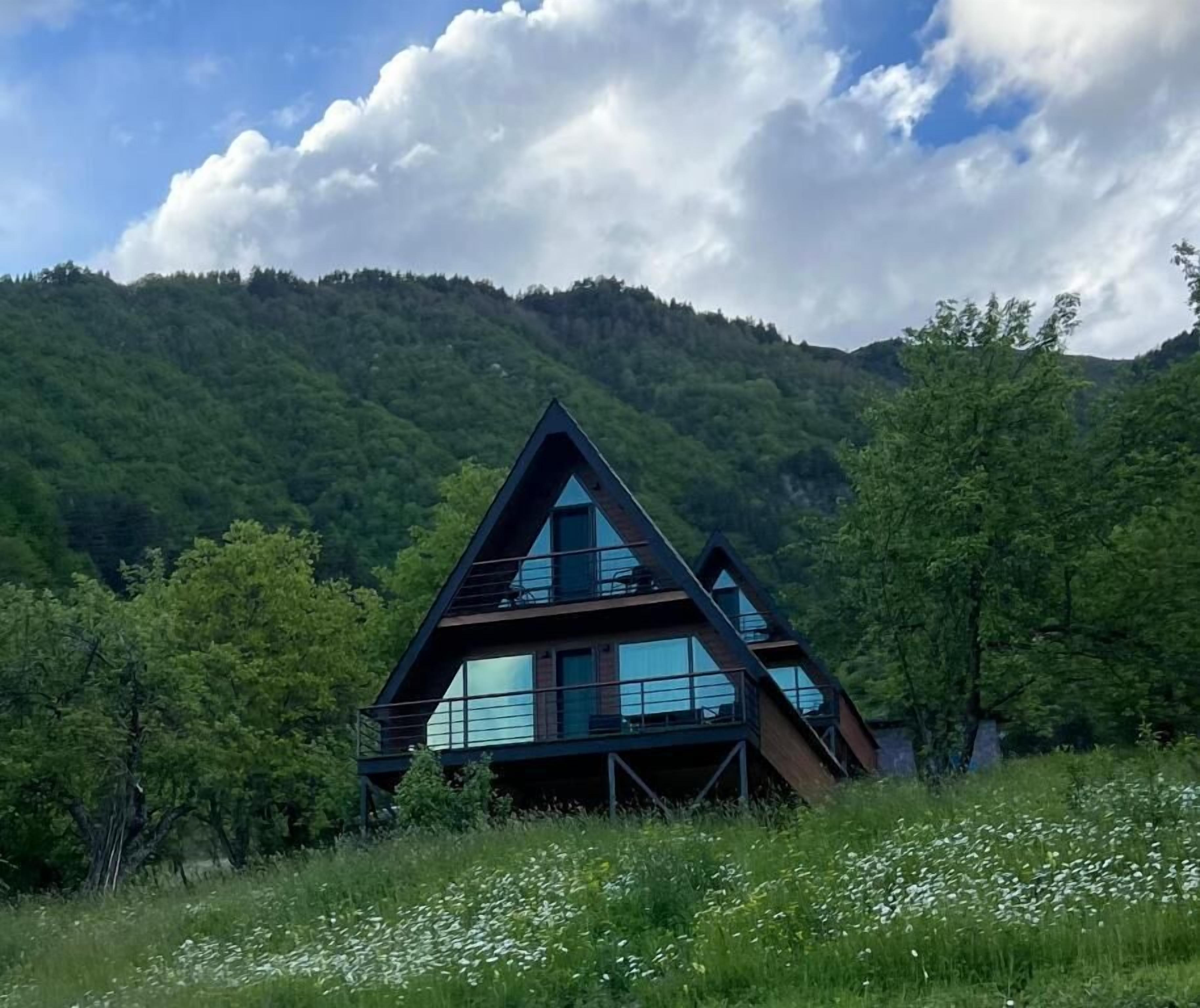 Un bâtiment moderne de forme triangulaire, doté de grandes fenêtres et entouré d’une végétation luxuriante et de montagnes.