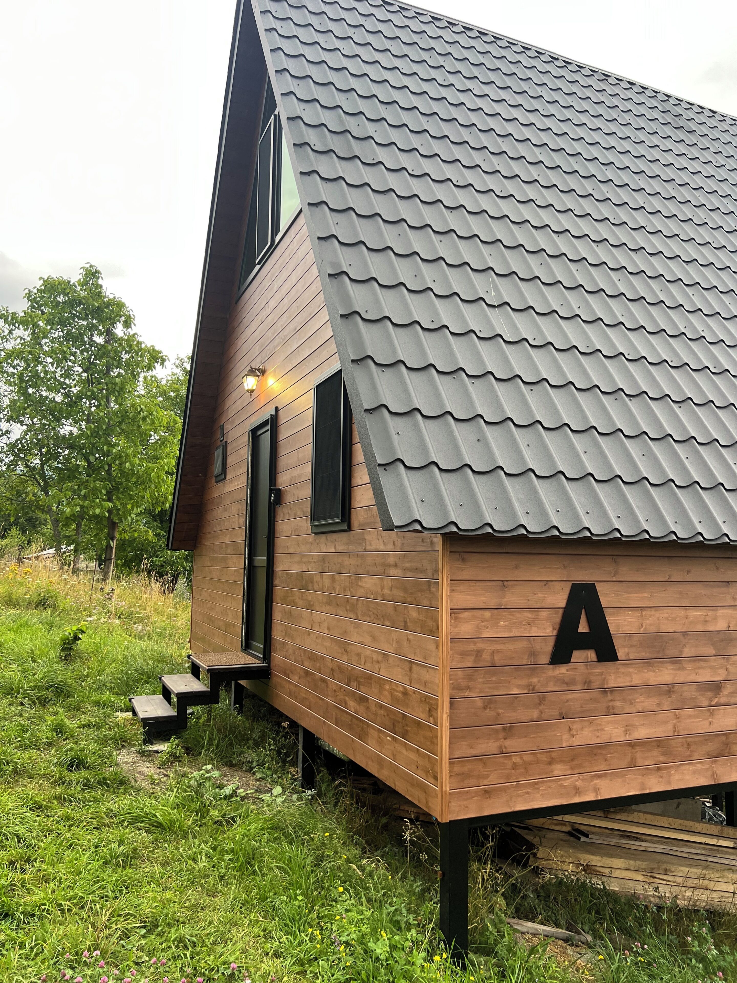 Een houten chalet met schuin dak, een letter 'A' aan de zijkant, en een klein terras met trap.