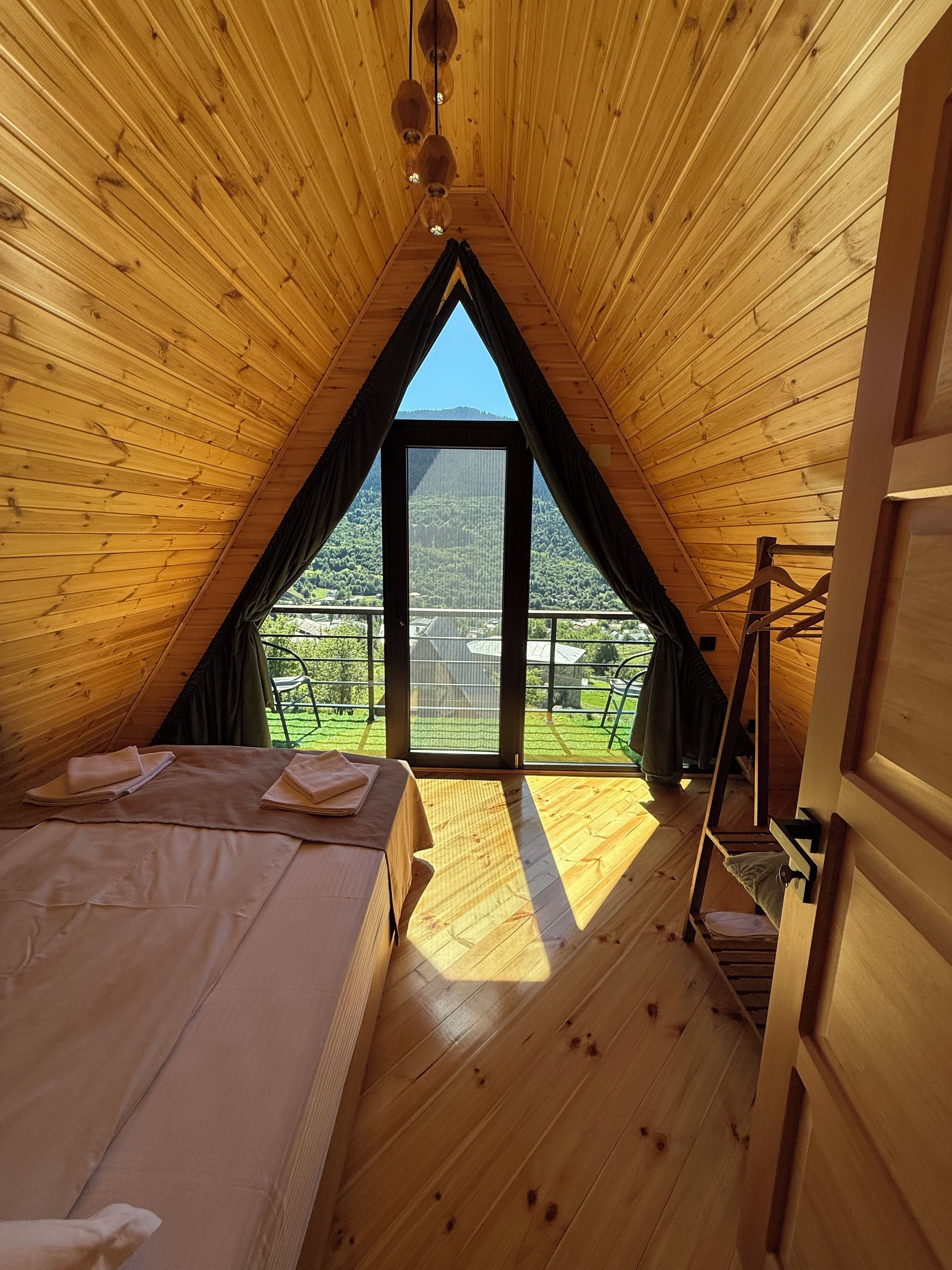 A wooden attic bedroom with a large window, a bed, and a ladder leading to a balcony.