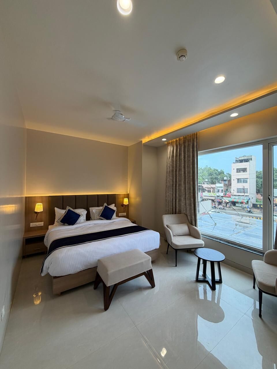 Habitación de hotel moderna con una cama grande, lámparas de noche, una mesita, una silla y vistas a la calle a través de un ventanal amplio.