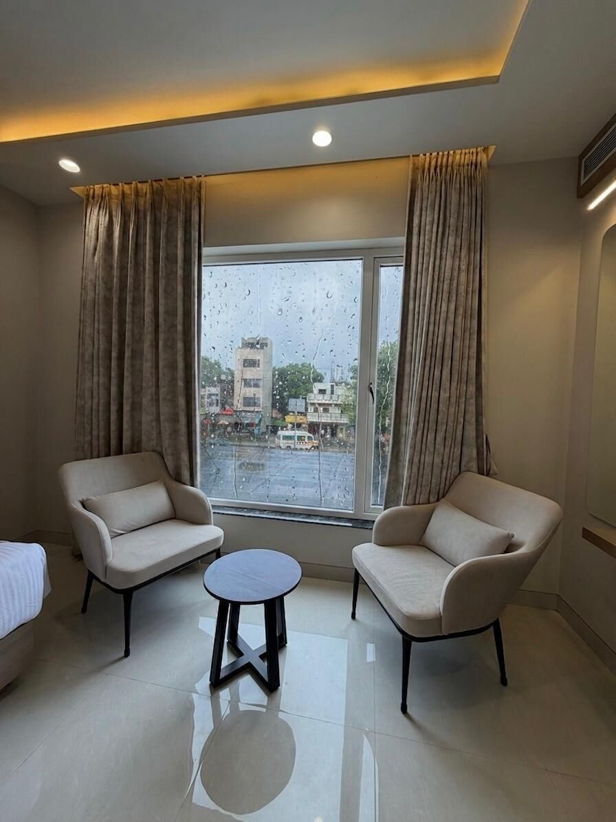 Ein Hotelzimmer mit zwei Sesseln, einem kleinen runden Tisch und einem großen Fenster mit Vorhängen.