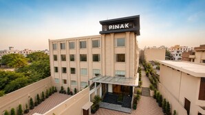Um edifício de vários andares com telhado plano, uma placa com a inscrição 'PINAK' e uma entrada central com marquise de vidro.