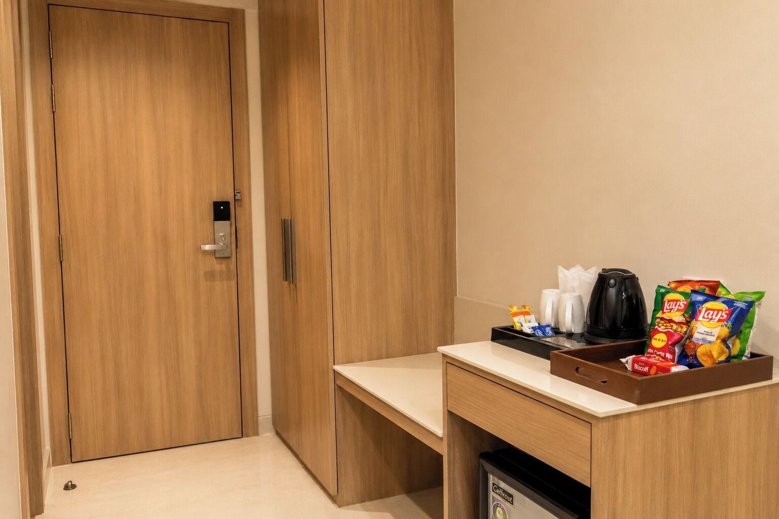 Habitación de hotel con un armario de madera, un pequeño escritorio con una bandeja de bocadillos, una cafetera y un cesto de basura.